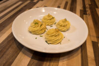 Four piped Potato Kisses Bramborové Pusinky topped with chives on a white plate on a wooden table.