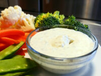 Ranch Dip Glass bowl of Ranch Dip surrounded by cauliflower, broccoli, carrots, and snap peas on a plate.