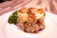 Rustic Crushed Potatoes with Bacon and Onion Square serving of Rustic Crushed Potatoes with Bacon and Onion, topped with crispy browned onion, on a white plate.