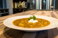 Czech Sauerkraut Soup Zelňačka A bowl of Czech Sauerkraut Soup Zelňačka with potato chunks, a dollop of sour cream, and a parsley garnish, set on a striped wooden countertop.