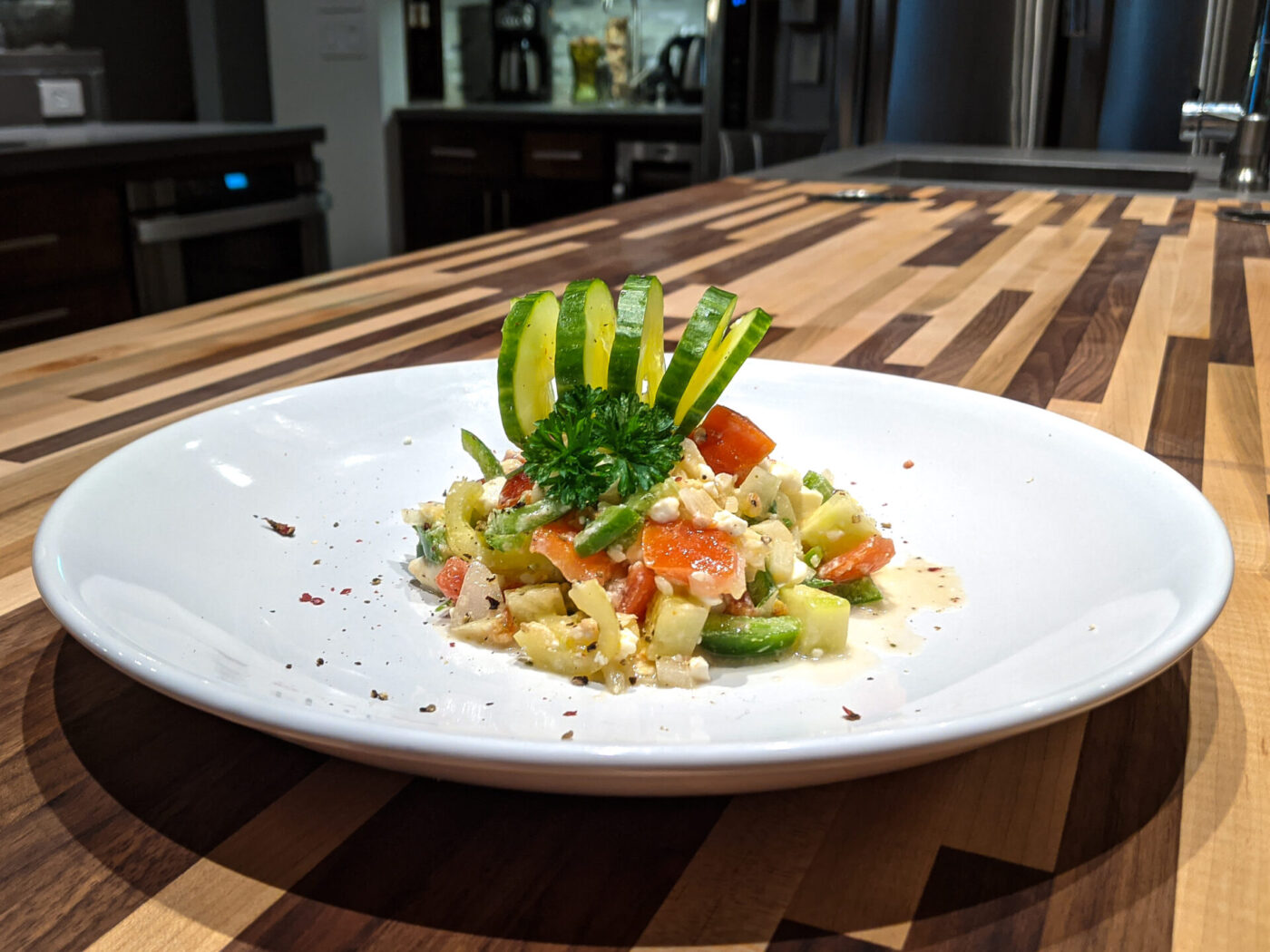 Diced Shopska Salad on a white plate, topped with a cucumber ribbon fan and herb sprig, on a striped wood counter.