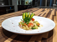 Diced Shopska Salad on a white plate, topped with a cucumber ribbon fan and herb sprig, on a striped wood counter.