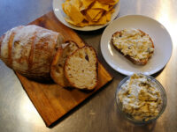 Artichoke Spinach Dip Glass bowl of Artichoke Spinach Dip beside a slice of bread spread with it, plus sourdough loaf and tortilla chips.
