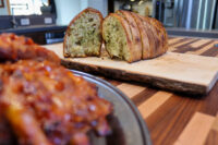 Twice-baked Herb Bread Sliced Twice-baked Herb Bread with green herb flecks on a wooden board beside glazed meat