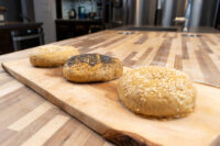 Bagel Recipe Three seed-topped bagels, two with sesame and one with poppy seeds, resting on a wooden board.
