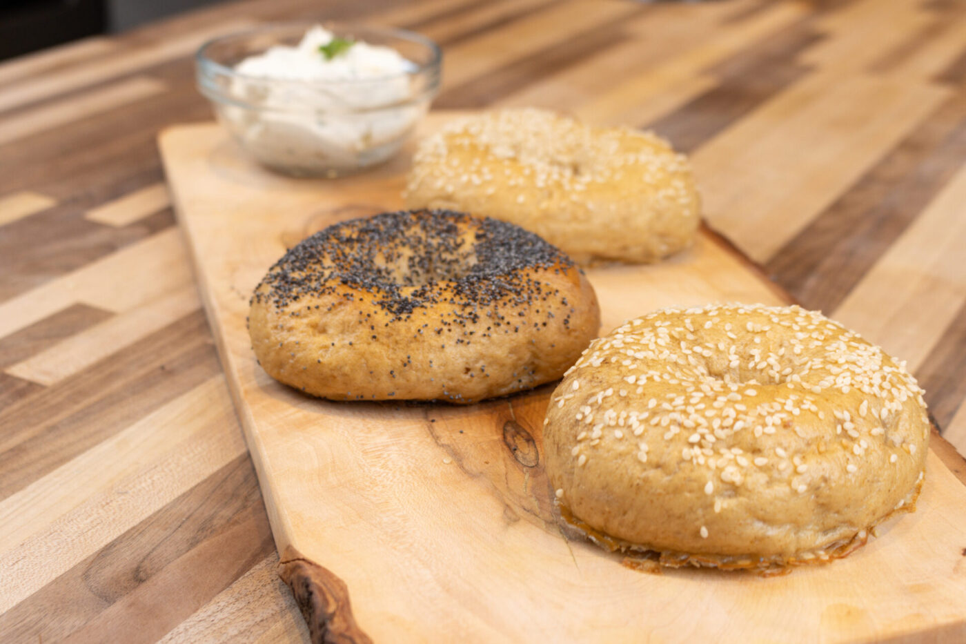 Bagel Recipe Three seeded Bagels, one topped with poppy seeds, two with sesame, on a wooden board beside a cream cheese dip.