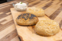 Bagel Recipe Three seeded Bagels, one topped with poppy seeds, two with sesame, on a wooden board beside a cream cheese dip.