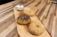 Bagel Recipe Three bagels, two sesame-topped and one poppy seed-topped, on a wood board beside a bowl of cream cheese