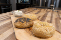 Bagel Recipe Three Bagels—poppy seed, sesame seed, and plain—on a wooden board beside a bowl of cream cheese.