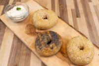 Bagel Recipe Three bagels, two with sesame seeds and one with poppy seeds, on a wooden board beside a bowl of cream cheese