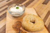 Bagel Recipe Sesame seed bagel on a wooden board beside a glass bowl of cream cheese with parsley.