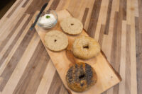 Bagel Recipe Wooden board with four sliced bagel halves, plain, sesame, and poppy seed, beside cream cheese.
