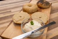 Creamy Spread with Garlic and Chives Glass bowl of Creamy Spread with Garlic and Chives, herb-topped, beside halved bagels and a knife on a wooden board.