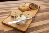 Creamy Spread with Garlic and Chives Assorted bagel halves on a wooden board next to a small bowl of Creamy Spread topped with a sprig of herb.