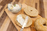 Bagel With Cream Cheese Halved bagel spread with cream cheese beside a whole sesame bagel and a bowl of cream cheese on a wooden board.