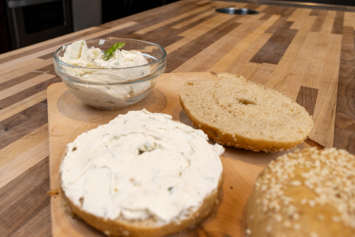 Bagel with Cream Cheese Bagel halves on a wooden board, one spread with herbed cream cheese, beside a bowl of the same spread.