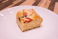 Bublanina (Czech Bubble Cake) Square slice of Bublanina (Czech Bubble Cake) with strawberries, dark berries, and powdered sugar on a white plate.