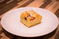 Bublanina (Czech Bubble Cake) Square slice of Bublanina (Czech Bubble Cake) with strawberry and berry pieces, dusted with powdered sugar, on a white plate.
