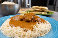 Butter Chicken on Rice Butter Chicken pieces in thick orange sauce over herbed rice on a blue plate, naan visible behind.