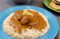 Butter Chicken Butter Chicken pieces in thick tomato sauce over herbed rice, garnished with citrus zest, on a teal plate.