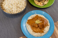 Butter Chicken Butter Chicken with pale garnish on rice, blue plate, beside rice pot and naan on green plate.