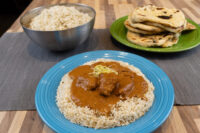 Butter Chicken Blue plate of Butter Chicken over rice, with a metal bowl of rice and flatbreads nearby.