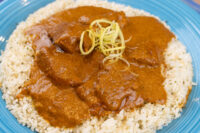 Butter Chicken Butter Chicken pieces in thick orange sauce over rice, topped with shredded garnish, on a blue plate.