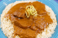 Butter Chicken Butter Chicken pieces in reddish-brown sauce over white rice, topped with thin julienned garnish, on a blue plate