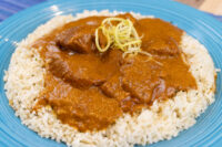 Butter Chicken Butter Chicken pieces in thick tomato sauce over herbed rice, garnished with citrus zest, on a blue plate.