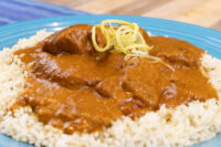 Butter Chicken Butter Chicken pieces in thick tomato sauce over rice, topped with shredded ginger, on a blue plate.