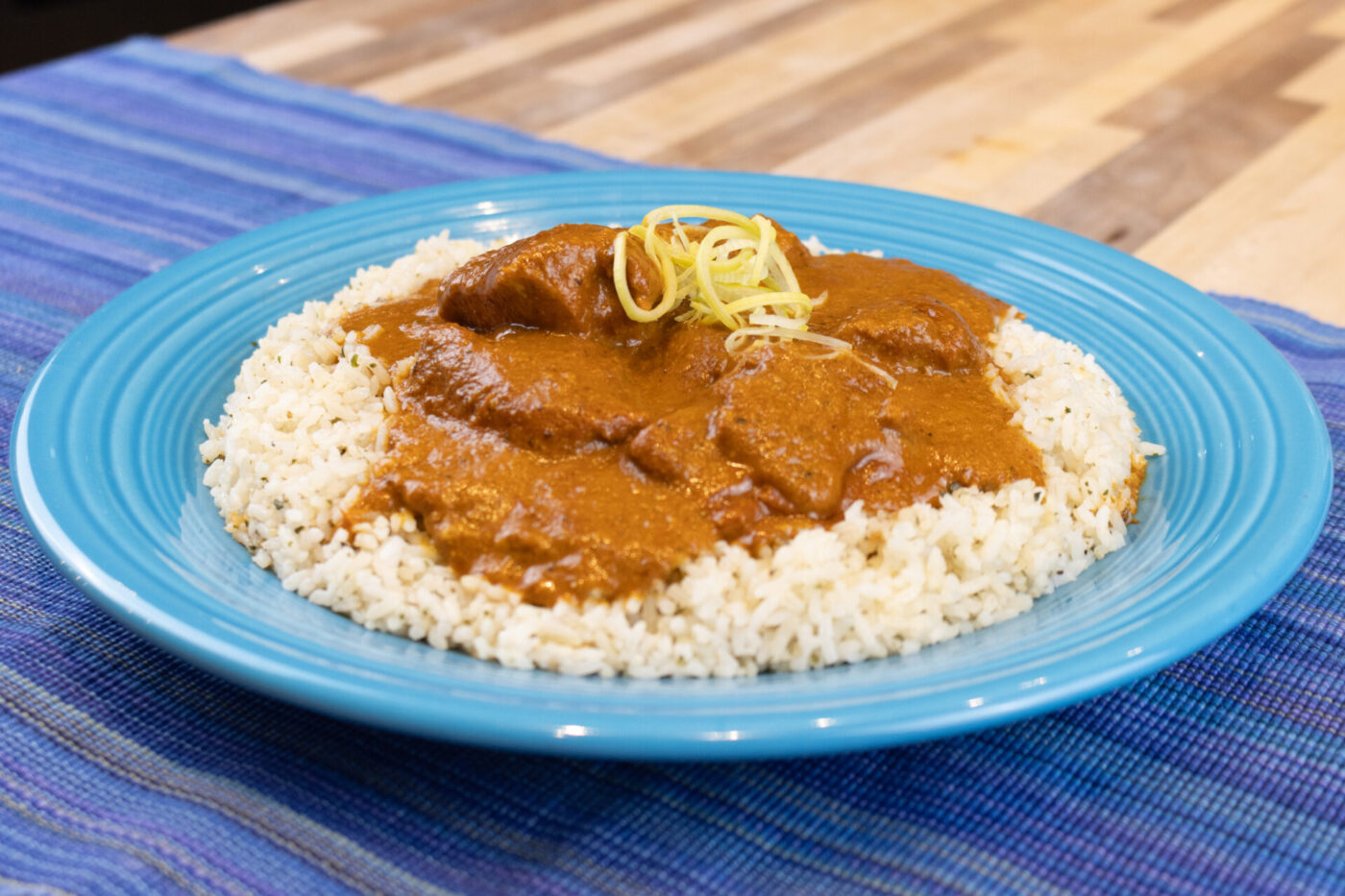 Butter Chicken Blue plate of Butter Chicken over rice, garnished with citrus zest curls, on a striped blue cloth