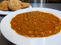 White bowl of Chana Masala, a thick orange-brown chickpea curry, with fried puri bread blurred behind.