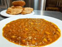 White bowl of Chana Masala chickpea curry with fried puri bread stacked on a plate behind.