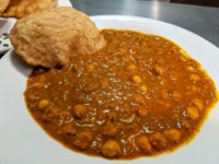 Chana Masala chickpeas in thick brown gravy on a white plate, served with puffed fried bread.