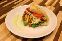 Chicken Gyros Plated Chicken Gyros sandwich with grilled chicken strips, lettuce, onion, and tomato in flatbread, plated with green sauce.