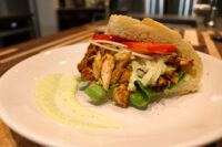 Chicken Gyros Chicken Gyros sandwich with sliced chicken, tomato, onion, spinach and green sauce in a roll on a white plate.