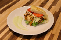 Chicken Gyros Chicken Gyros sandwich with grilled sliced chicken, lettuce, tomato and onion in a roll, green sauce swirl on plate.