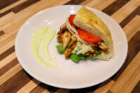Chicken Gyros Chicken Gyros in flatbread with lettuce, onion, and tomato, served with a green herb sauce on a white plate.