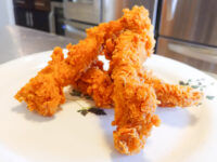 Chicken Tenders Four crispy Chicken Tenders stacked on a white plate with a small herb sprig garnish.