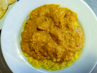 Chicken Tikka Masala Chicken Tikka Masala in a white bowl, its creamy orange sauce covering turmeric-yellow rice, with a piece of flatbread beside it