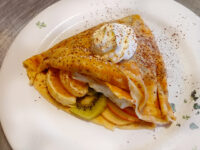 Crepes Folded Crepes filled with banana, orange, kiwi and apple slices, topped with whipped cream and a dusting of spice on a white plate.