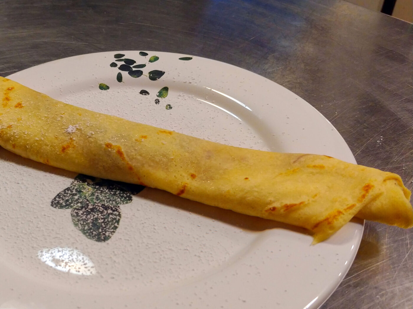 Crepes A rolled Crepes with a lightly speckled golden surface rests diagonally on a white patterned plate.