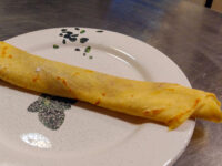 Crepes A rolled Crepes with a lightly speckled golden surface rests diagonally on a white patterned plate.