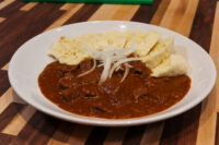 A plate of Czech Beef Goulash with tender beef chunks in dark paprika sauce, topped with raw onion slices and bread.