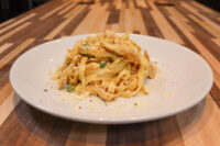 Fettuccine Alfredo with Chicken Fettuccine Alfredo with Chicken piled on a white plate, showing cream-coated ribbon pasta, chicken strips, peas, and grated cheese on a striped wooden table.