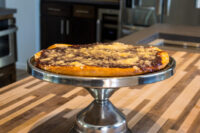 Whole round Frgal Kolach with Plum Filling, streusel-topped, on a metal pedestal stand on a wood counter.