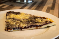 Triangular slice of Frgal Kolach with Plum Filling topped with streusel, on a white plate.