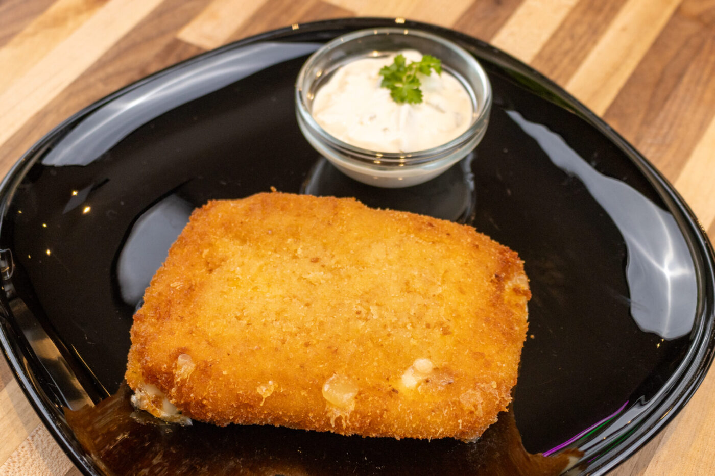 Fried Cheese with Ham Golden breaded Fried Cheese cutlet on a black plate beside a small dish of herbed white dip.