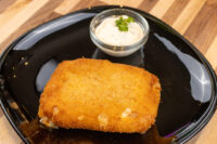 Fried Cheese with Ham Golden breaded Fried Cheese cutlet on a black plate beside a small dish of herbed white dip.