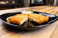 Fried Cheese with Ham Fried Cheese with Ham cut in half showing melted cheese and ham, served with a small dip on a black plate.